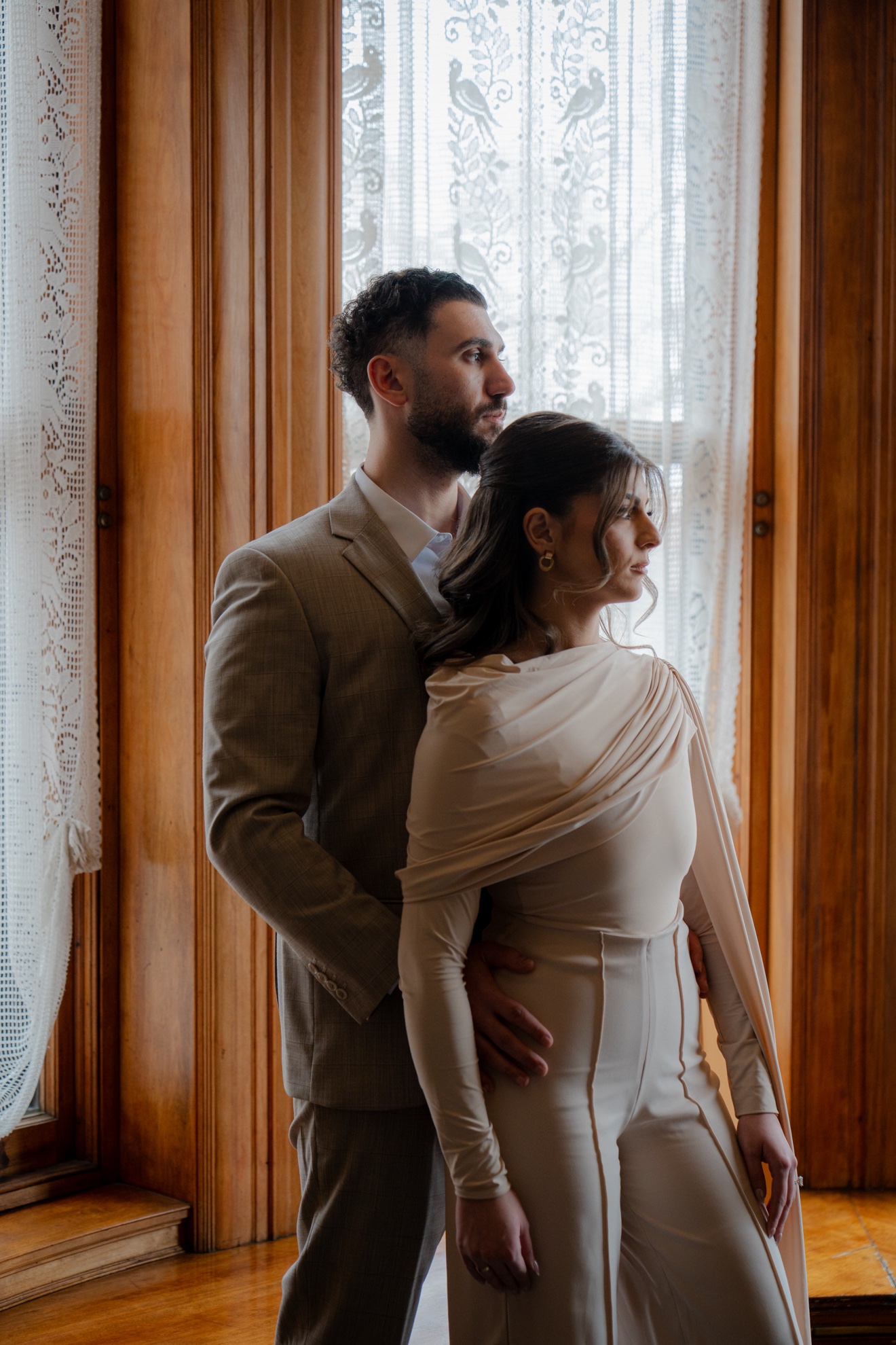 Hussein and Selena standing together beside a lace-curtained window